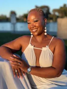 Erica "EMissyK" sits outdoors near a calm lake during golden hour, wearing a light sleeveless dress and white spherical earrings. She poses gracefully in warm sunlight with trees and greenery softly blurred in the background.